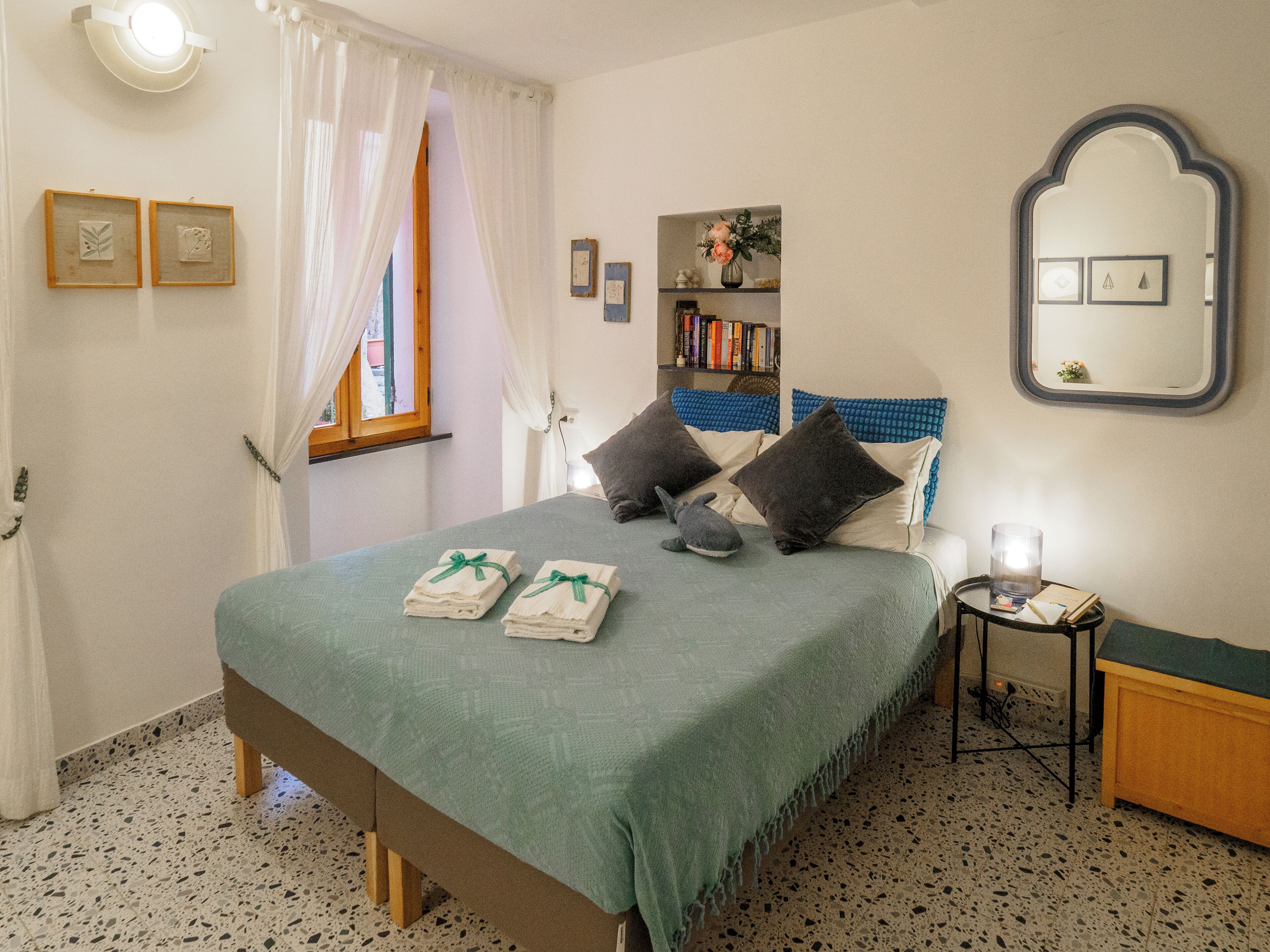 A neatly made bed with a green bedspread, two pillows, and a stuffed shark toy. There are two framed pictures on the wall, a bookshelf with books, and a mirror with a decorative frame.