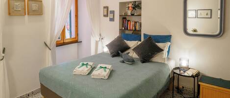 A neatly made bed with a green bedspread, two pillows, and a stuffed shark toy. There are two framed pictures on the wall, a bookshelf with books, and a mirror with a decorative frame.