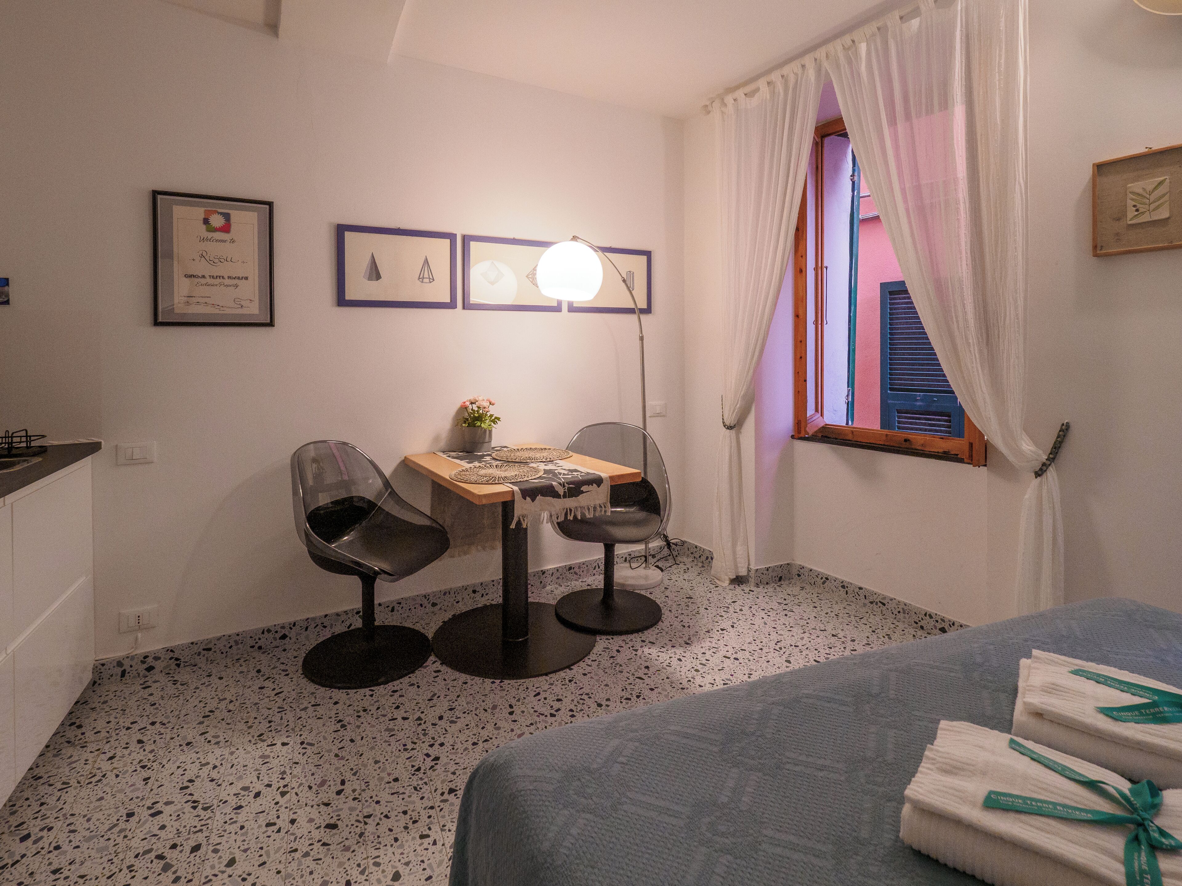 A small, neatly arranged room with a bed, a table with chairs, a desk with a lamp, and a window with curtains.