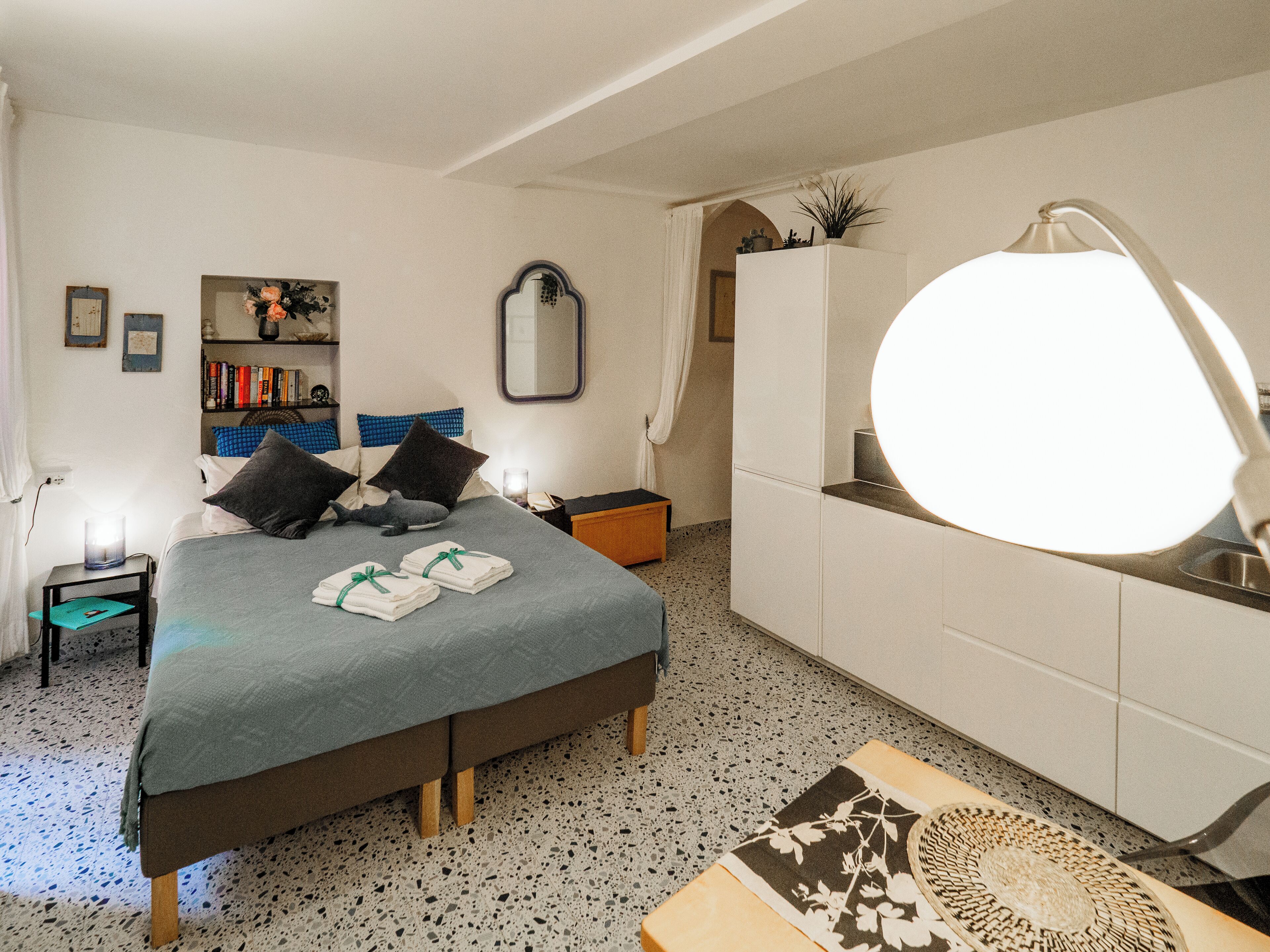 Un dormitorio con cama, mesita de noche, cómoda, espejo y una pequeña zona de lectura con sillón y mesa.