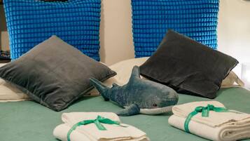 A bed with blue and gray pillows, a blue shark plush toy, and neatly folded white towels with green ribbons.