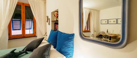 A bedroom with a bed, a window with curtains, a mirror, and a small table with a bottle.