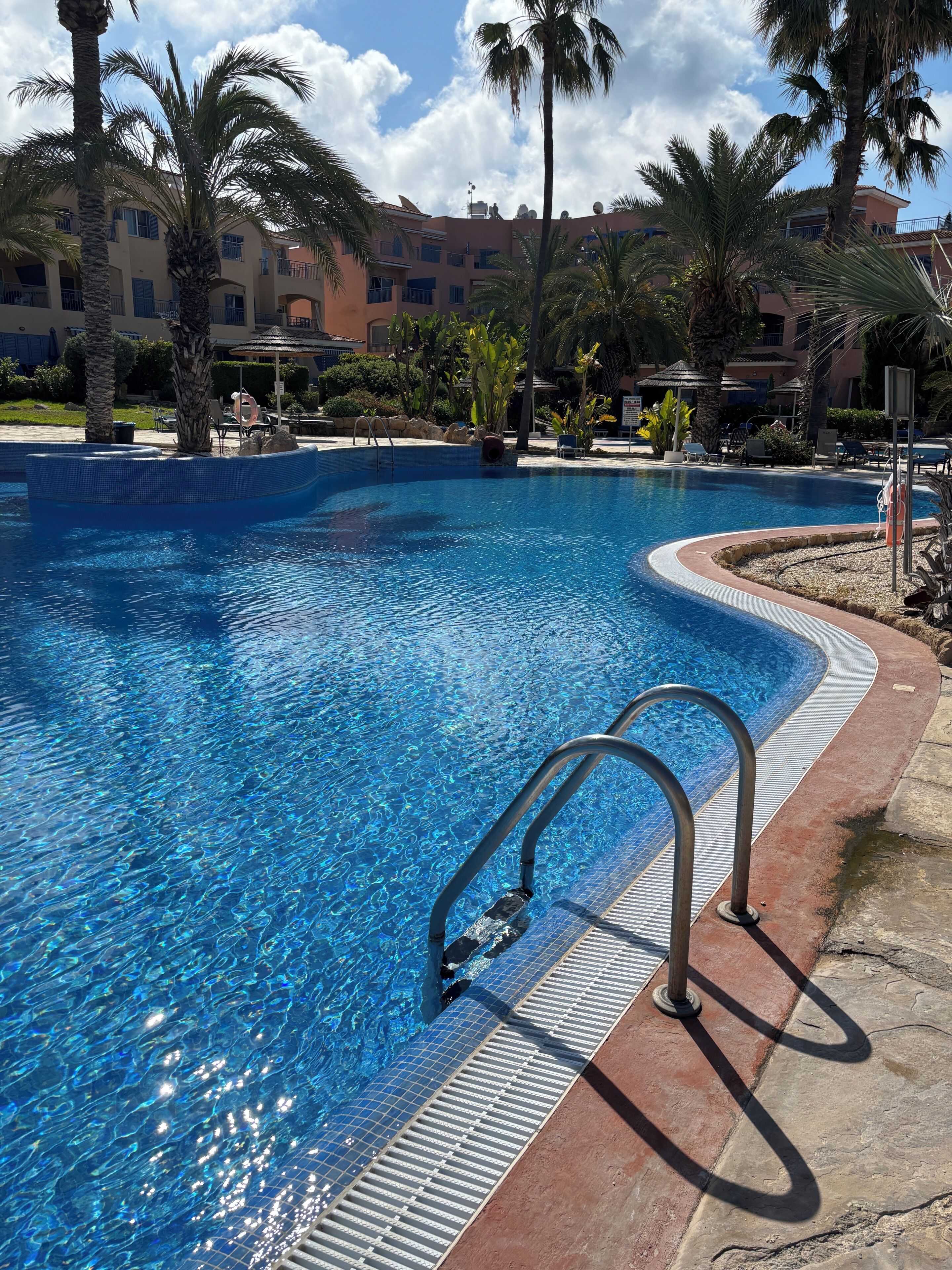 A swimming pool with clear blue water, surrounded by palm trees and a building with balconies.