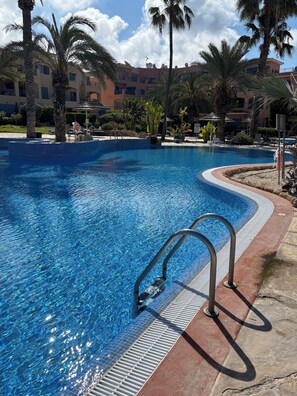 Une piscine à l’eau d’un bleu cristallin, entourée de palmiers et bordée d’un bâtiment doté de balcons.