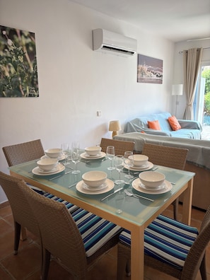 Ein Essbereich mit einem Glastisch, gedeckt für sechs Personen, Weidenstühle, ein Bett mit blauer Bettdecke und orangenen Kissen sowie eine Klimaanlage an der Wand.
