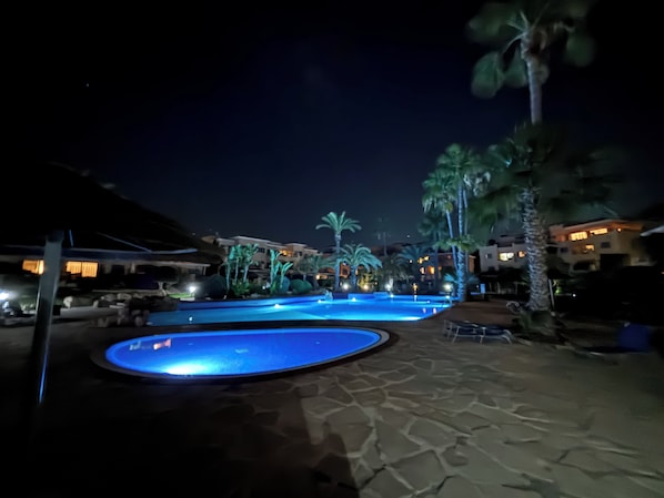 Ein Poolbereich bei Nacht mit einer Steindeck, Palmen und beleuchteten Gebäuden im Hintergrund.