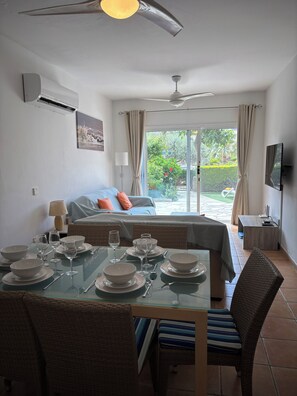 A room with a dining table set for four, a bed with a view, a ceiling fan, and a TV on the wall. - Family-Friendly Ground Floor Apartment with Garden, BBQ & Pools Limnaria Gardens (Kato Paphos)