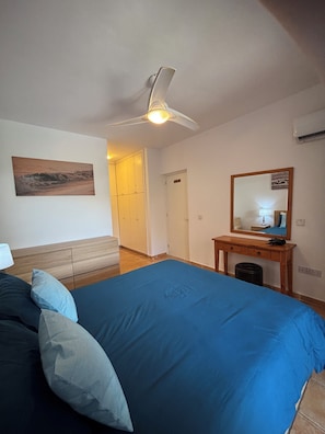 Ein Schlafzimmer mit einem großen Bett, einem Deckenventilator, einem Nachttisch mit Lampe, einem Spiegel und einer Tür zu einem Badezimmer.