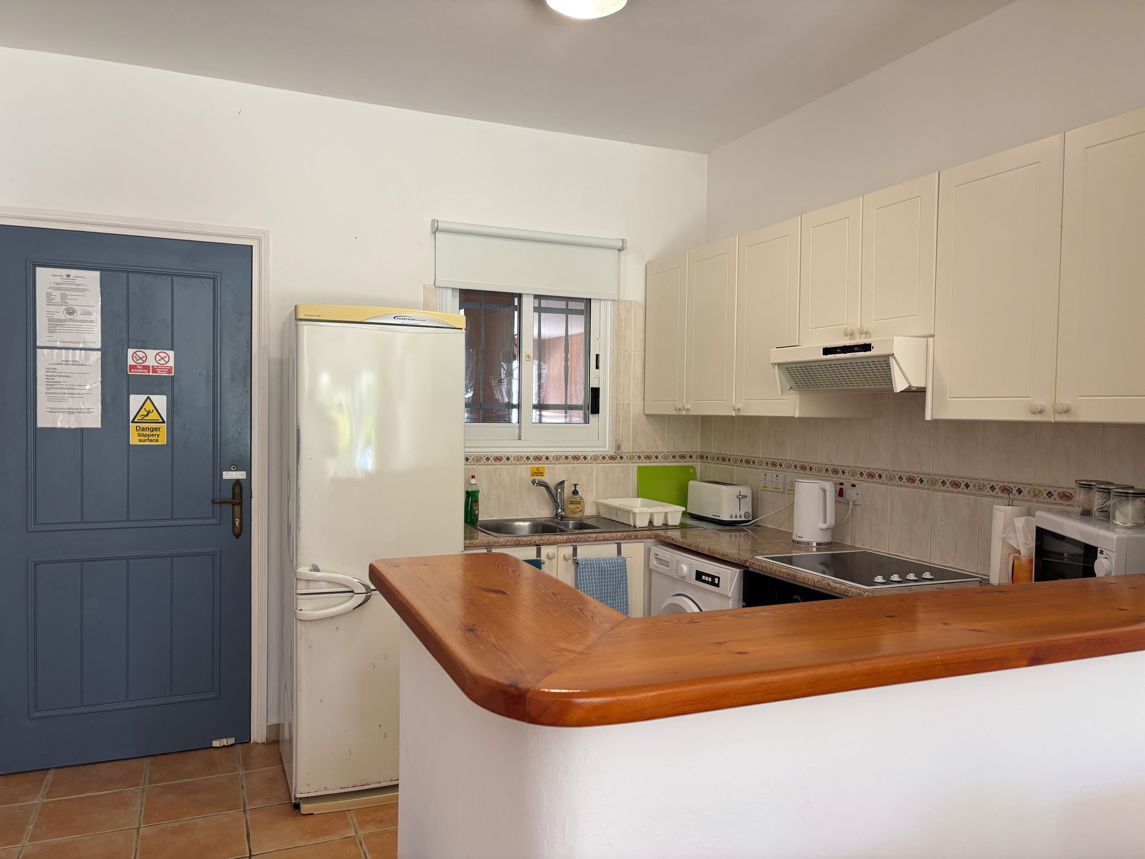 Eine Küche mit blauer Tür, weißen Schränken, einer Holz Arbeitsplatte, einem Kühlschrank, einer Waschmaschine und einem Spülbecken.