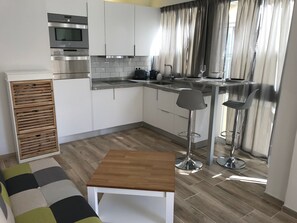 Una cocina moderna con armarios blancos, electrodomésticos de acero inoxidable, un mueble de madera con cajones, una barra con taburetes y una pequeña mesa.