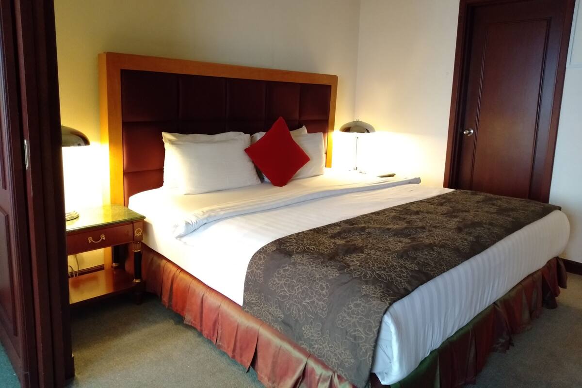 A neatly made bed with a wooden headboard, a red pillow, and a patterned bedspread. There is a bedside table with a lamp and a small table with a lamp. A wooden door is visible in the background.