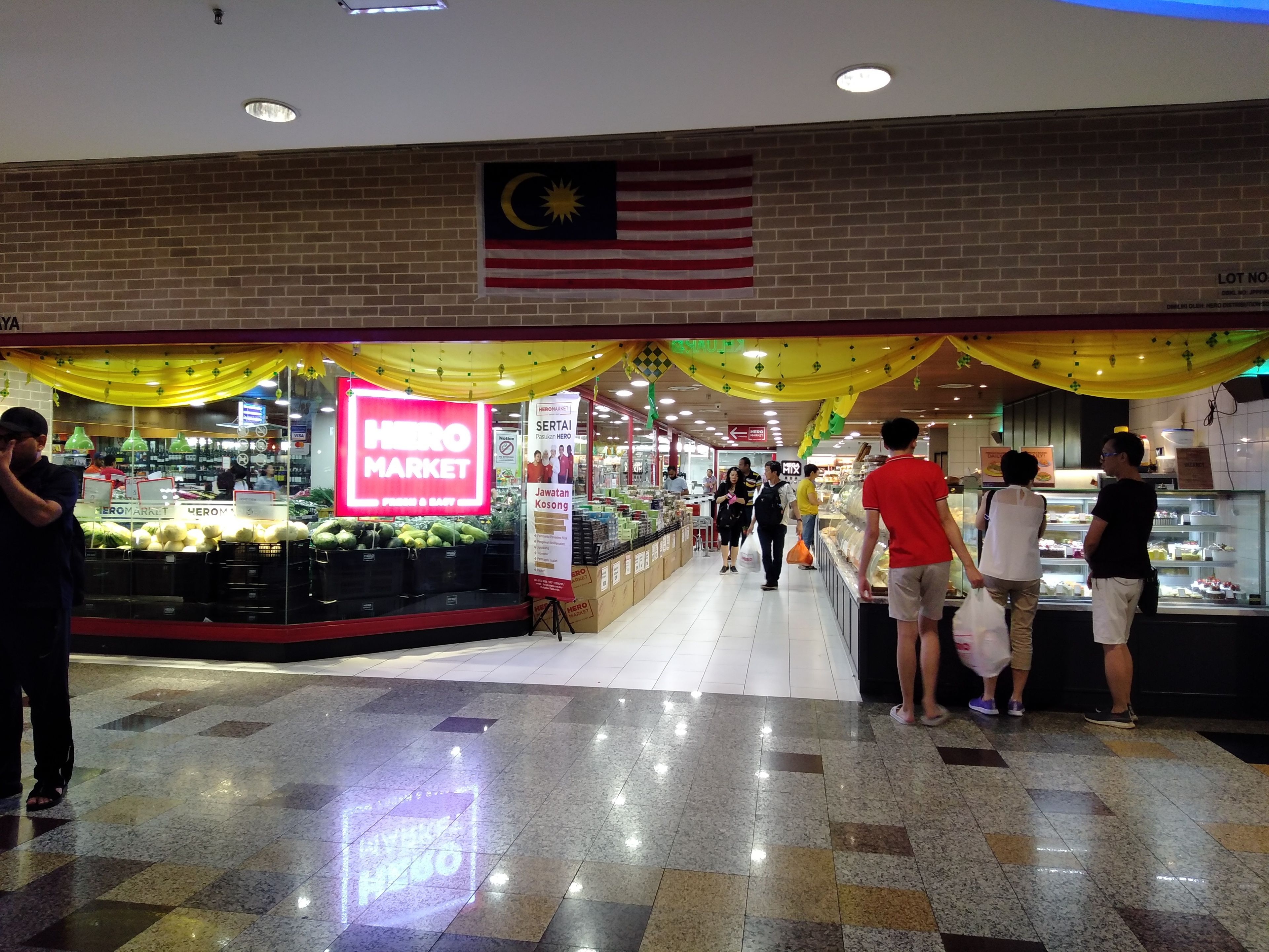 Um mercado com uma placa de destaque HERO MARKET, uma bandeira malaia e piso de azulejos.