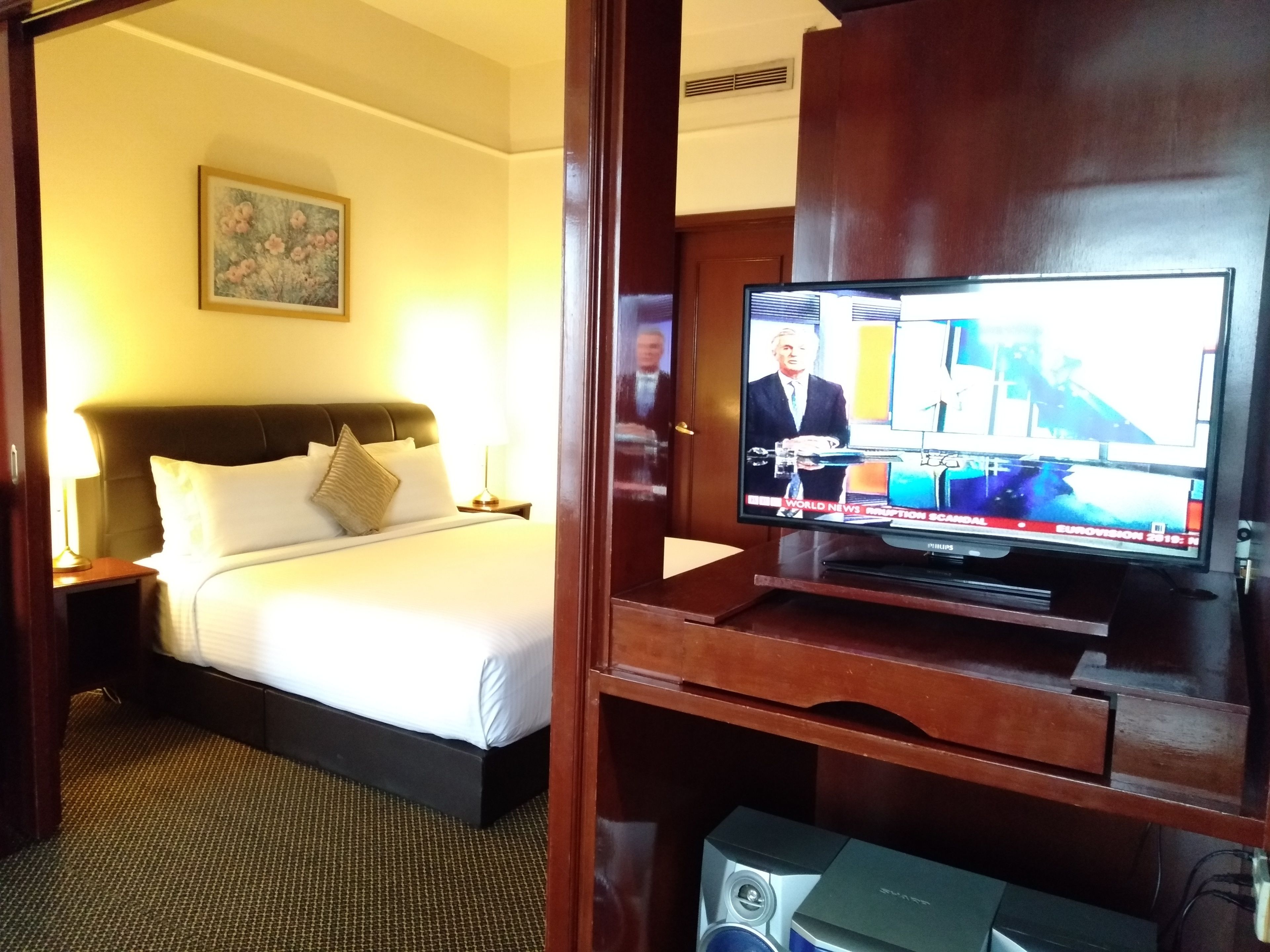 A hotel room with a bed, a television displaying a news channel, and a wooden cabinet.