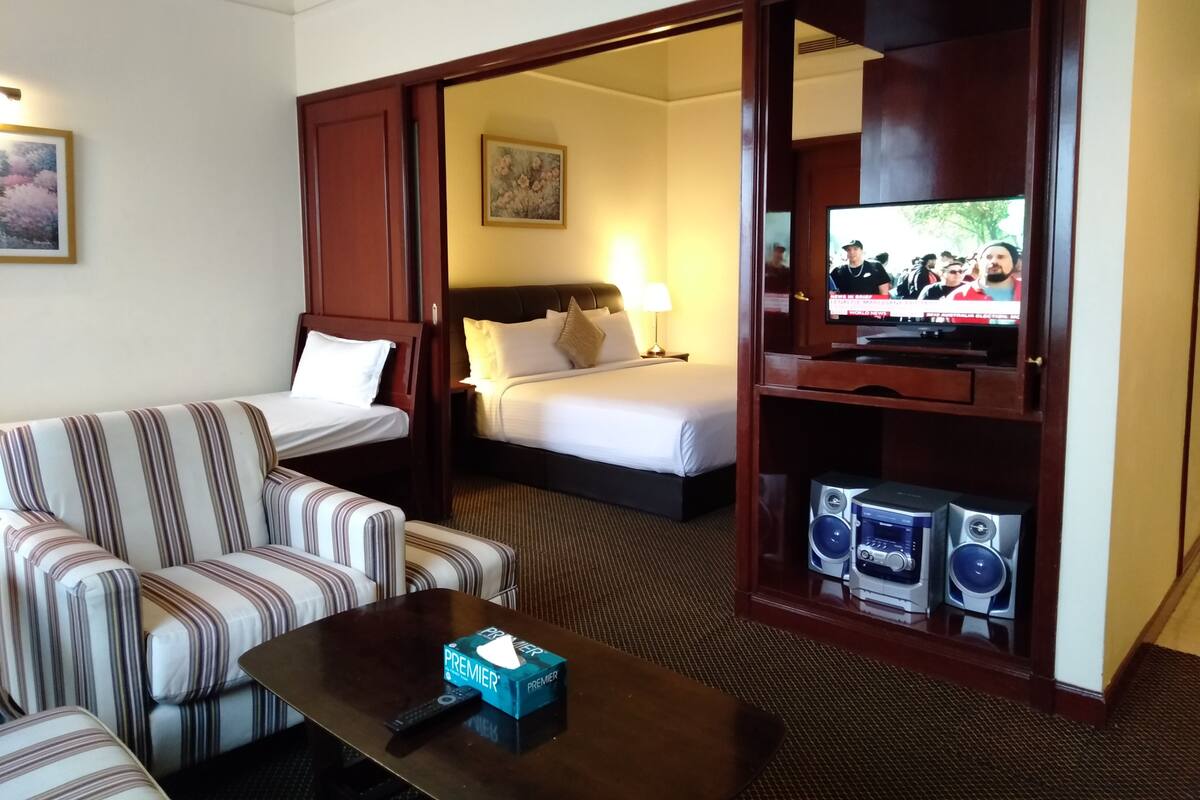 A hotel room with a striped sofa, a bed with white bedding, a television on a wooden stand, and a minibar.