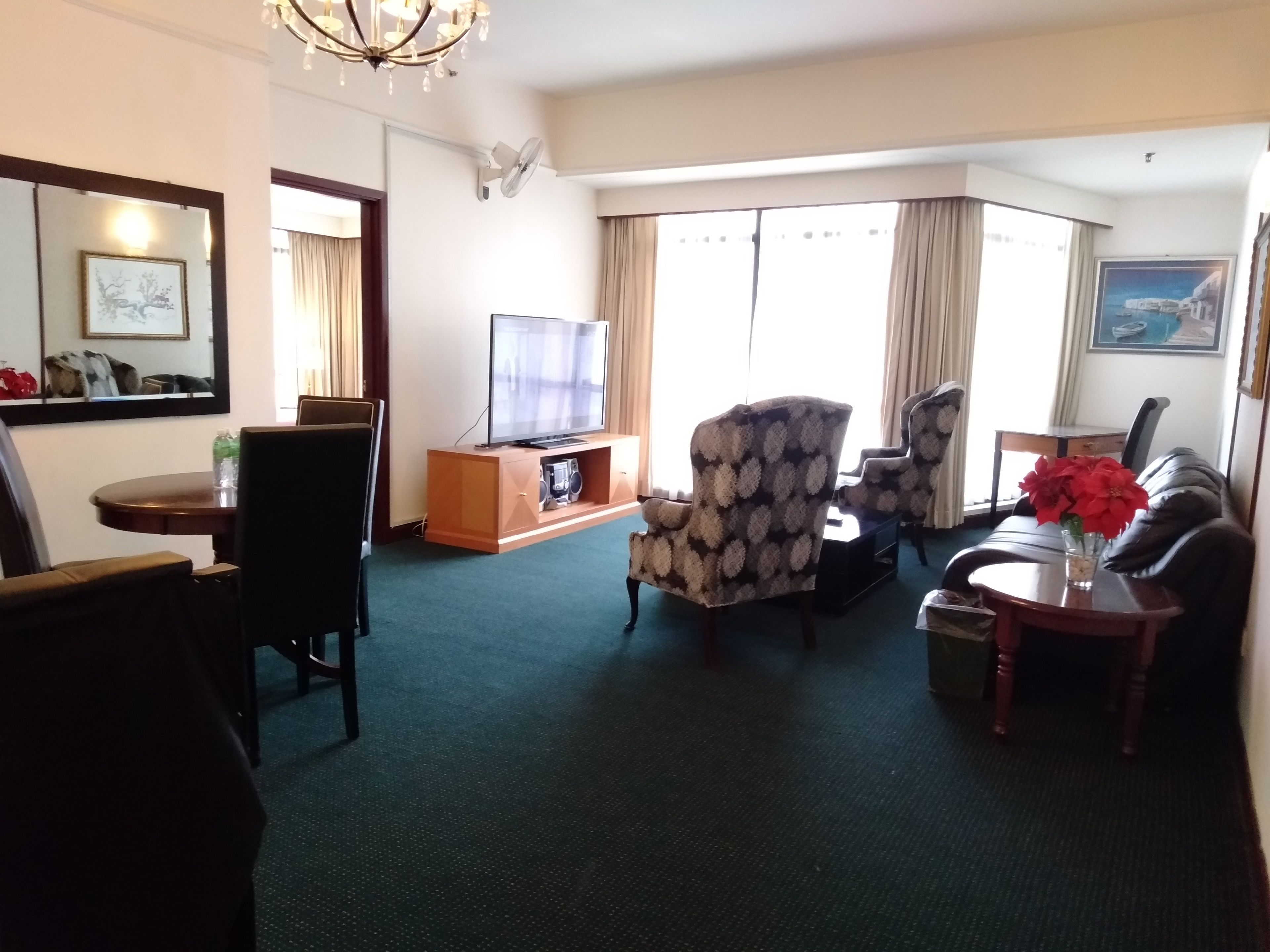 A hotel living room with a flat-screen TV, two patterned armchairs, a sofa, a coffee table, and a small side table with a vase of red flowers.