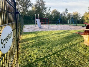 Children can play on the playground's slide and climbing frames, surrounded by a grassy area and trees.