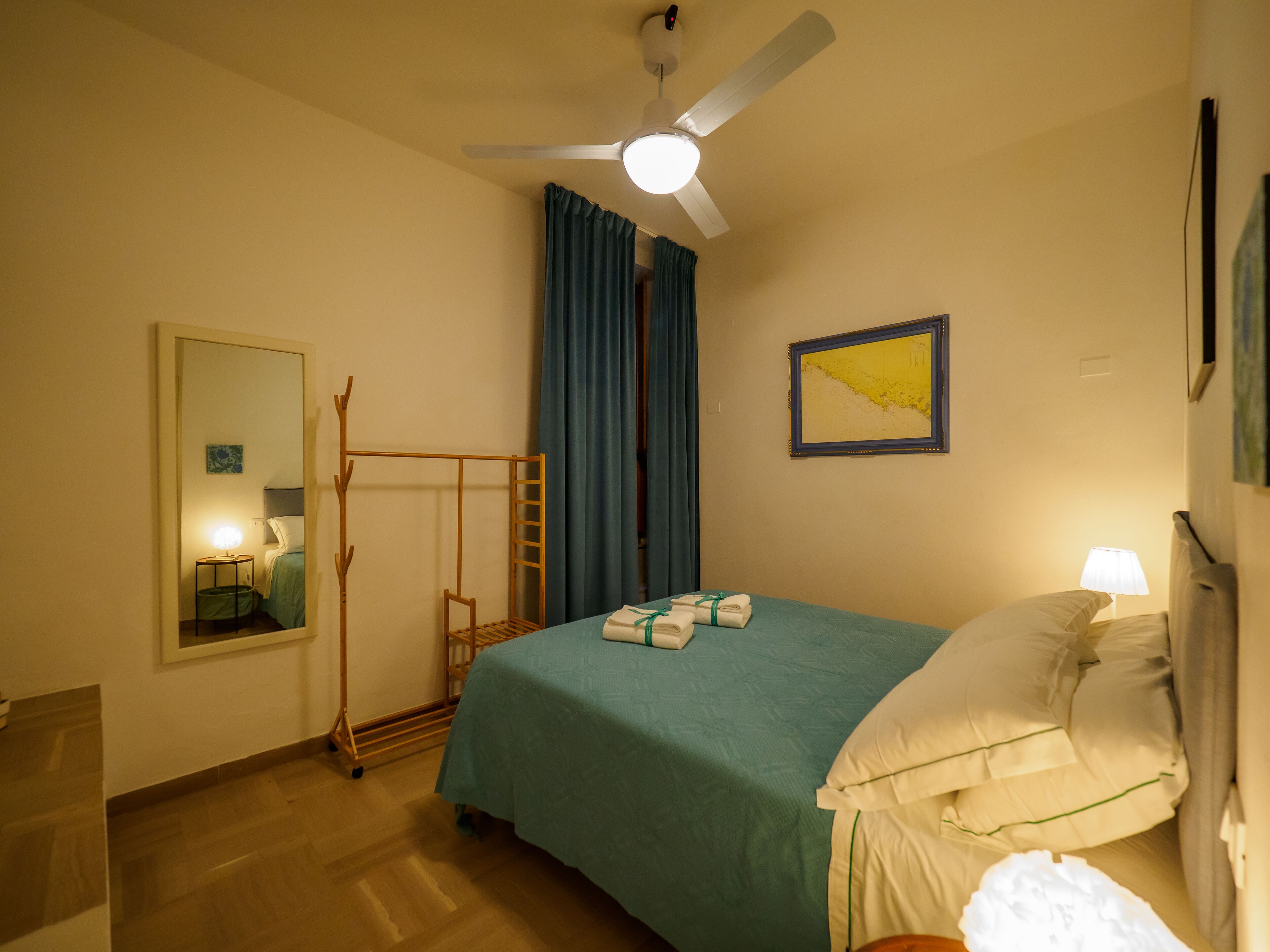 Habitación de hotel con cama, ventilador de techo, mesita de noche con lámpara, espejo, escalera de madera y un cuadro en la pared.