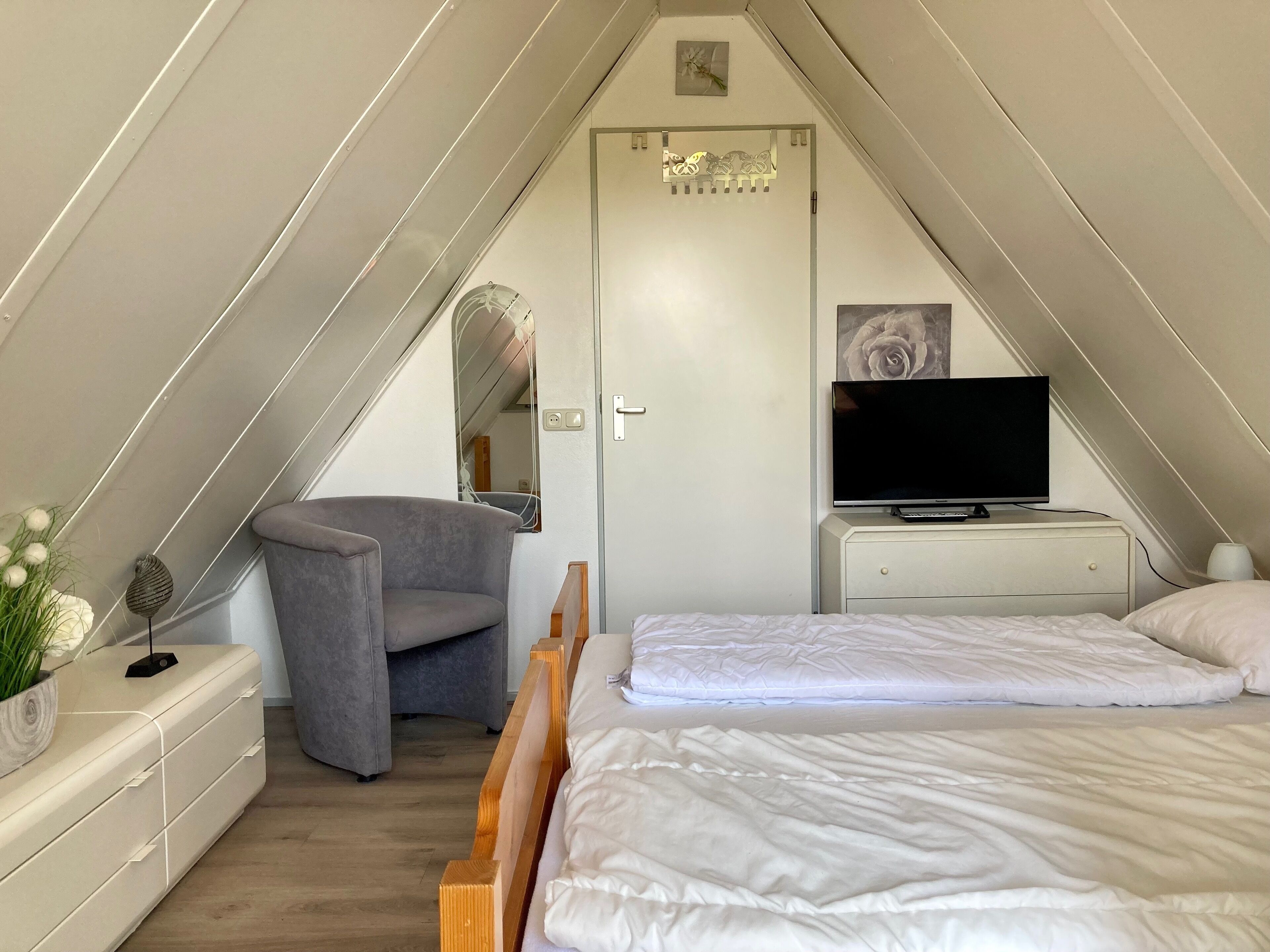 A bedroom with a sloped ceiling, a bed, a chair, a dresser, a television, and a door with a decorative panel.
