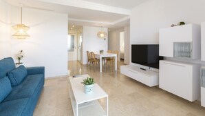 A modern living room with a blue sofa, a coffee table with a plant, a dining area with a table and chairs, a TV on a white cabinet, and a hallway leading to another room. - New Condo in the best Resort, Modern decoration - only families (Playa de Gandía)