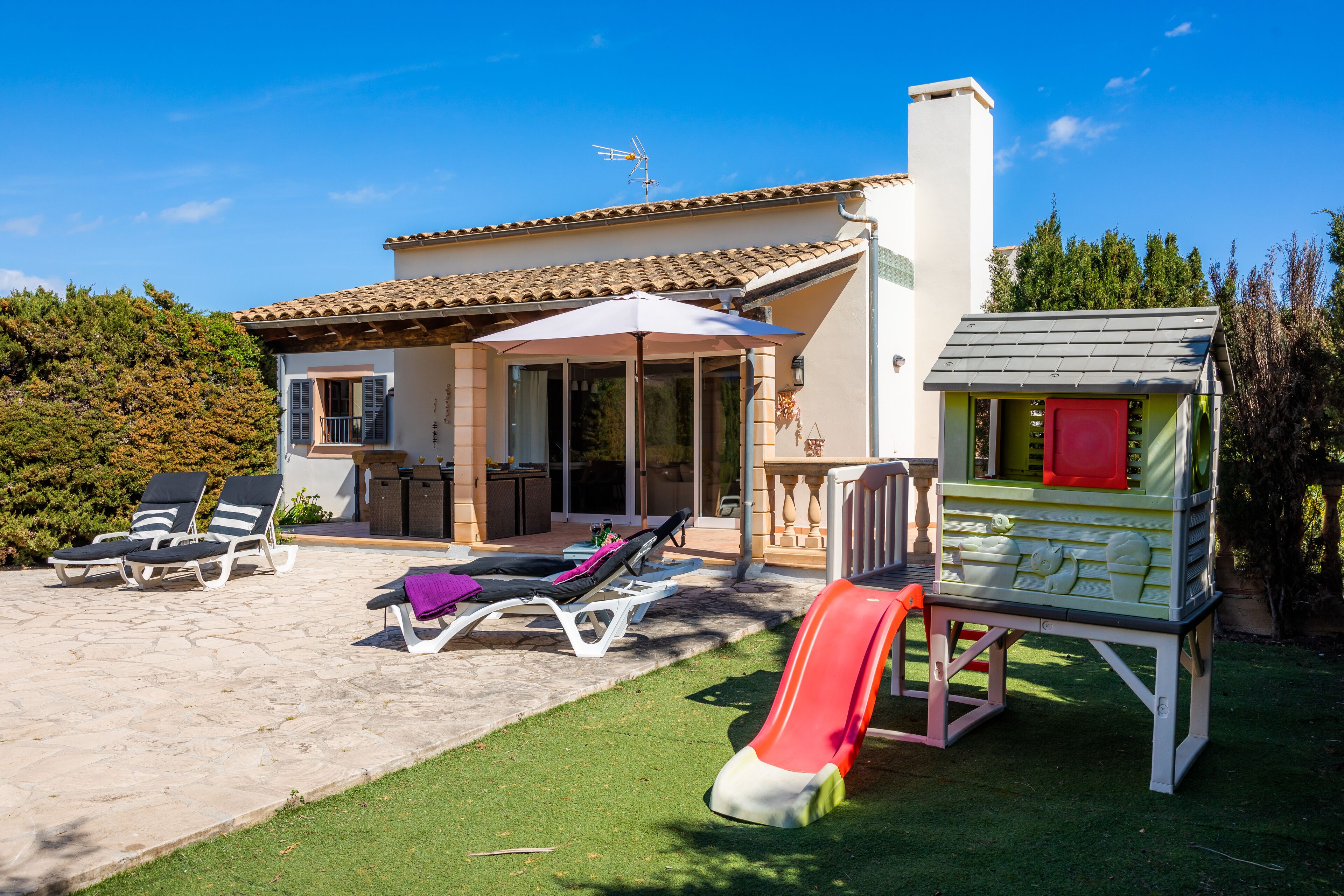 Een huis met een terras, buitenmeubels, een speelhuisje en een glijbaan.
