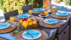 Un espace de restauration en plein air, avec des chaises et des tables en osier, où sont disposés des assiettes, des couverts et des verres de jus d’orange.