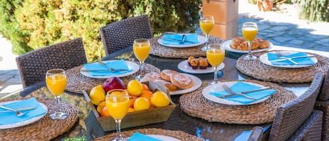 An outdoor dining area with wicker chairs and tables, set with plates, cutlery, and glasses of orange juice.
