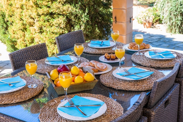 An outdoor dining area with wicker chairs and tables, set with plates, cutlery, and glasses of orange juice.