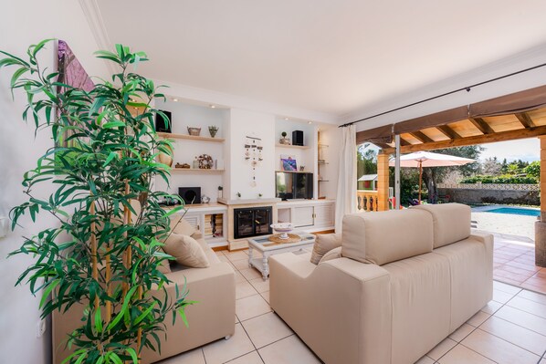 A spacious living room with a beige sofa, a coffee table, and a large bamboo plant. There is a fireplace and a TV on a shelf. The room has a large glass door leading to an outdoor area with a pool.