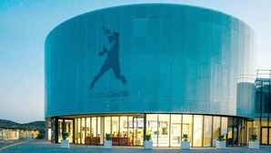 Un centre sportif moderne doté d’une grande façade en verre et d’une silhouette marquante représentant un joueur de tennis.