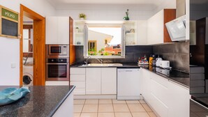 Une cuisine moderne avec des meubles blancs, un micro-ondes encastré, un four et un lave-vaisselle. Une fenêtre offre une vue sur l’extérieur, et une porte en bois donne accès à une autre pièce.