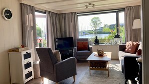A living room with a TV, a sofa, a chair, a small table, and a view of a lake with boats. - Chalet Gaby with sauna, private dock, and canoes at Lauwersmeer (Anjum)