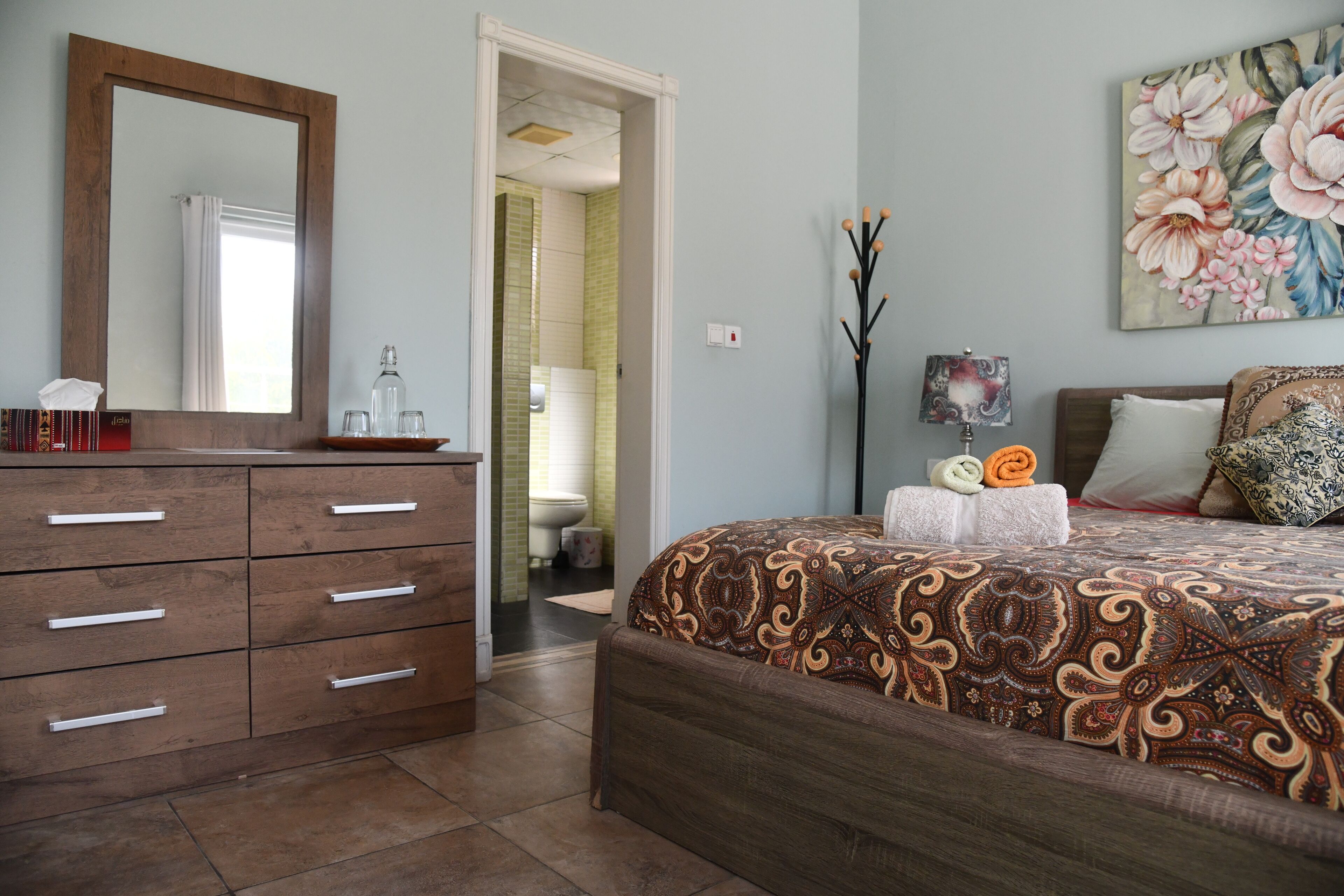 A bedroom with a wooden dresser, a large bed with patterned bedding, a painting on the wall, and a bathroom visible through an open door.
