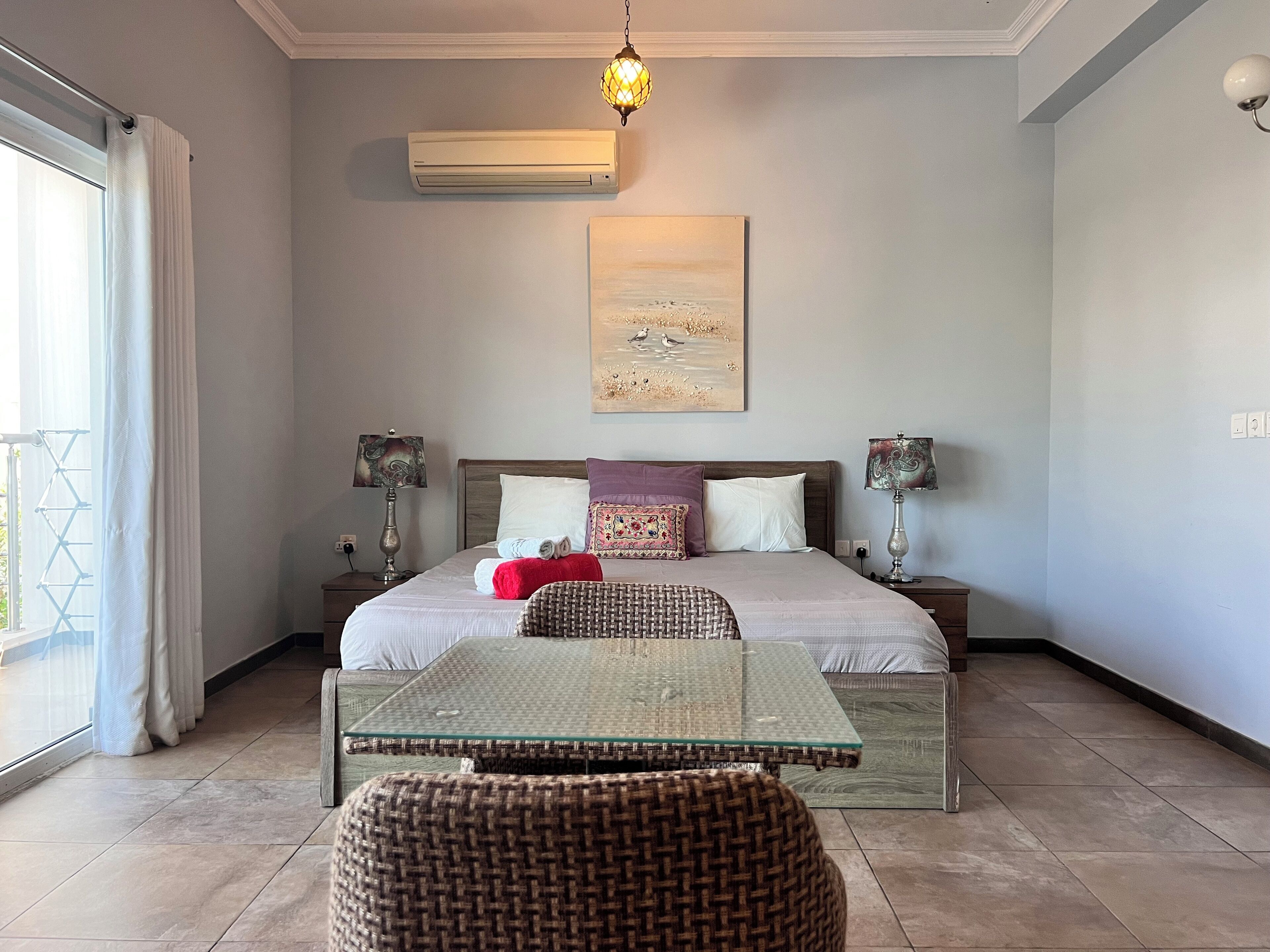 Un dormitorio con una cama, dos mesitas de noche, una mesa de centro de vidrio, una silla de mimbre, un aire acondicionado instalado en la pared y un cuadro.