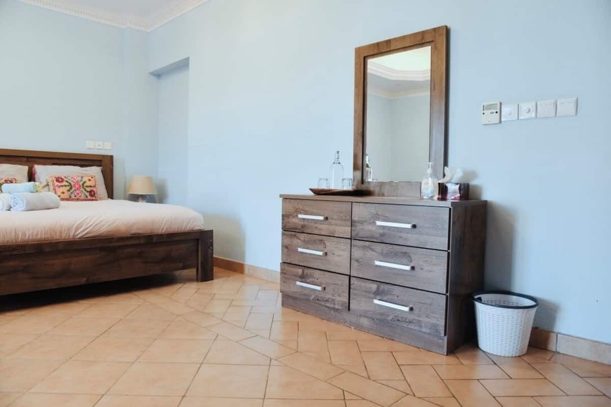 A bedroom with a bed, a wooden dresser with drawers, a mirror, and a waste basket.