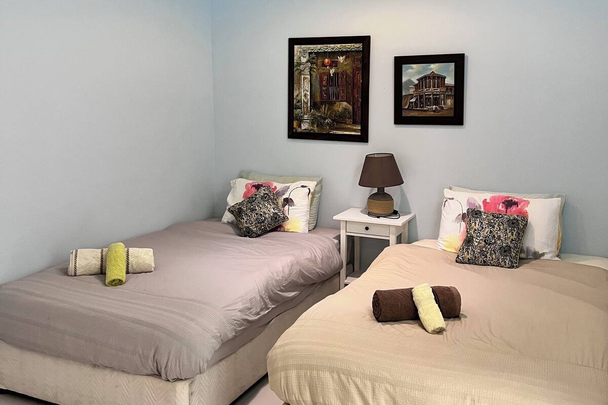 A hotel room with two beds, a ceiling fan, an air conditioner, a nightstand with a lamp, and two framed pictures on the wall.