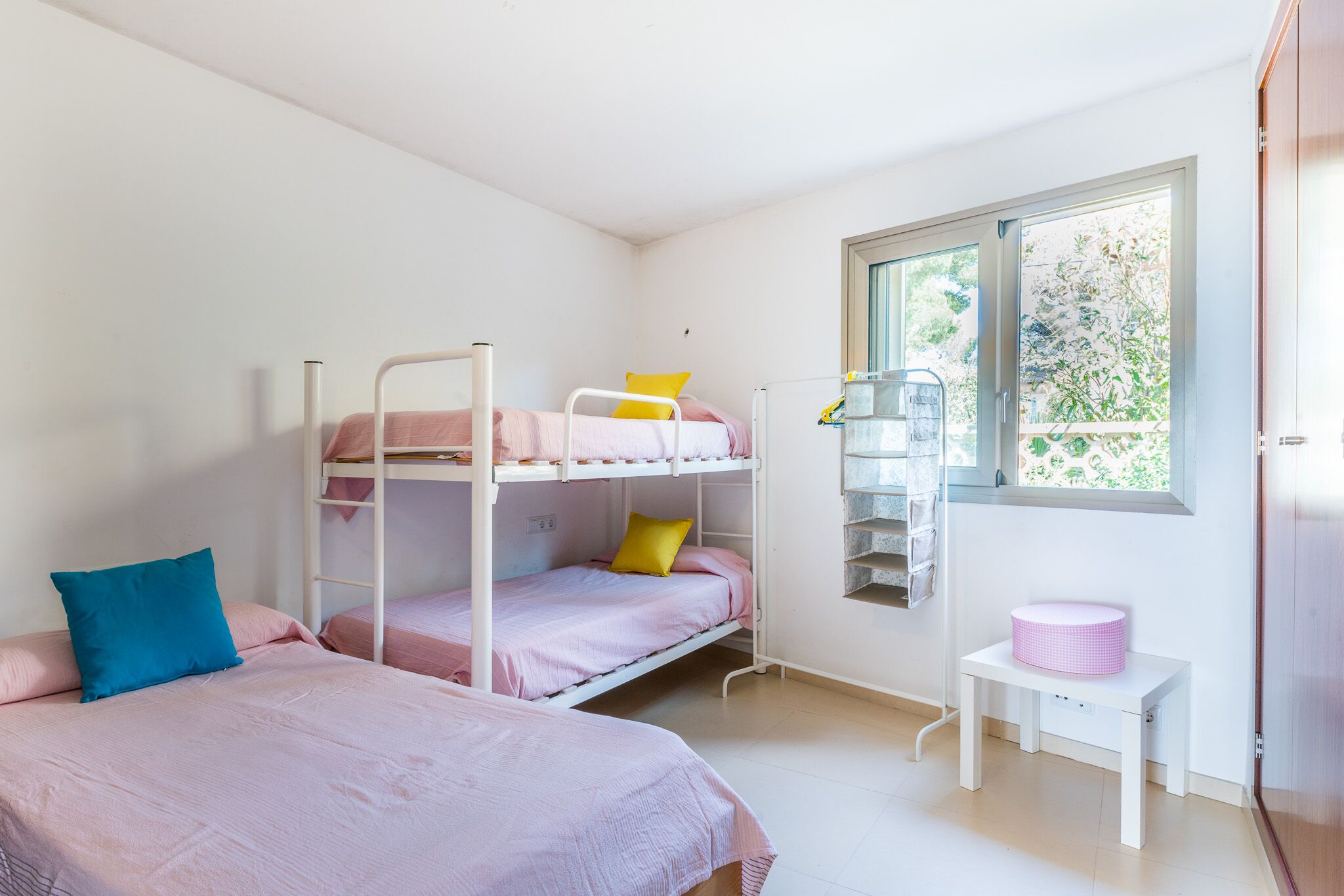 A dormitory room with bunk beds, a small table with a pink basket, and a window with a view of greenery.