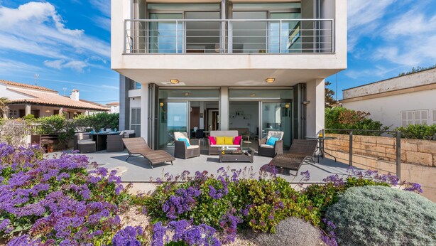 A modern two-story house with a balcony, outdoor seating area, and a garden with purple flowers.