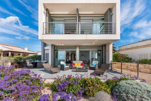 Une maison moderne à deux étages, dotée d’un balcon, d’un espace extérieur aménagé et d’un jardin où poussent des fleurs violettes.
