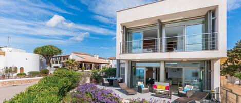 A modern two-story house with a balcony, outdoor seating area, and a garden with purple flowers.