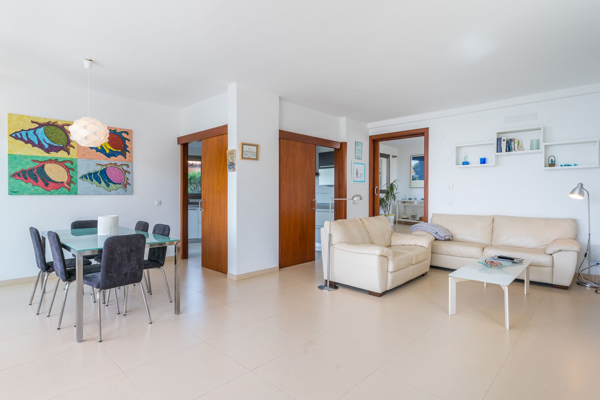 A modern living room with a glass dining table, black chairs, a beige sofa, and a white coffee table. There is a colorful artwork on the wall and a wooden door leading to another room.