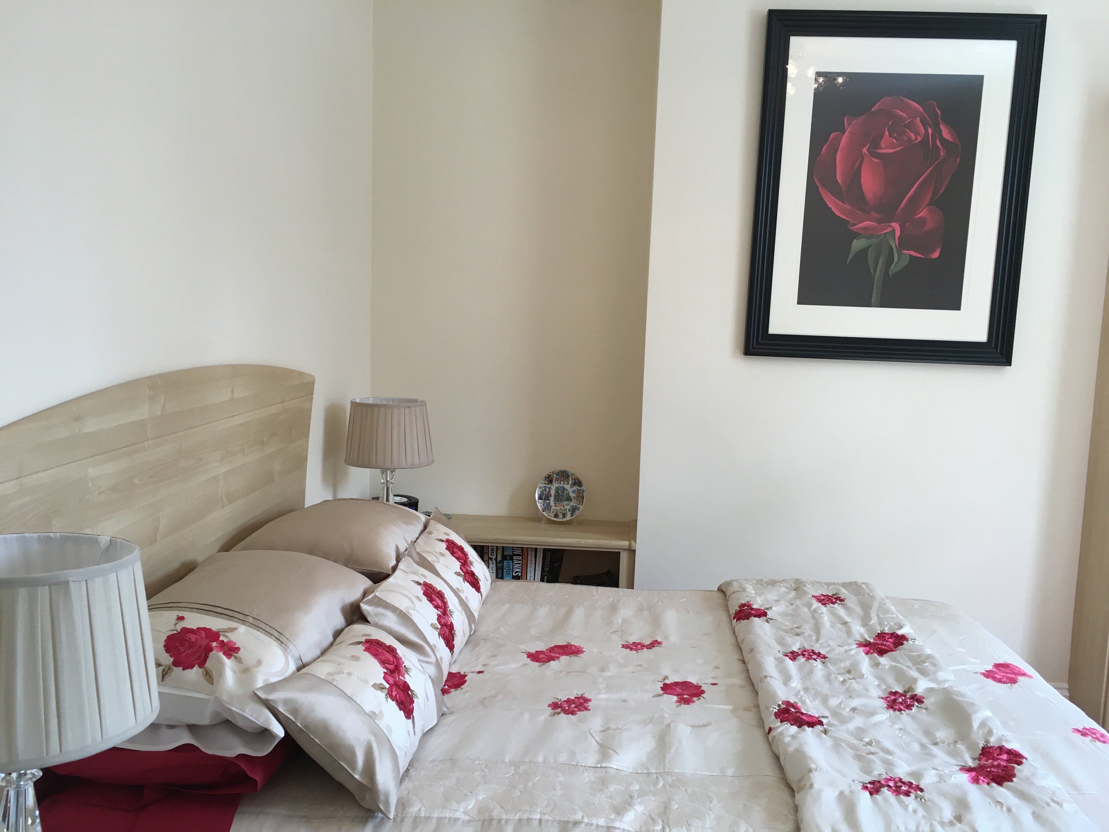 A neatly arranged bedroom with a bed, pillows, a bedside lamp, a framed picture on the wall, and a small table with a decorative plate.