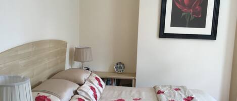 A neatly arranged bedroom with a bed, pillows, a bedside lamp, a framed picture on the wall, and a small table with a decorative plate.