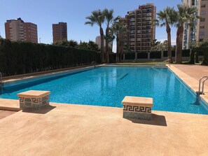 A swimming pool with two stone pedestals, surrounded by palm trees and tall buildings in the background. - APARTMENT IN PONIENTE BEACH R033 (Benidorm)