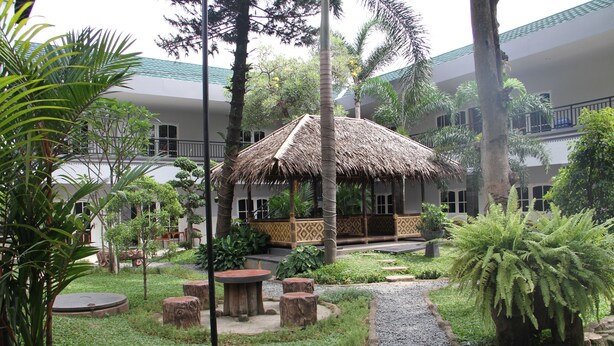 A courtyard with a thatched-roof structure, surrounded by palm trees and ferns, with a pathway leading to a building with balconies. - Mandala Hotel (Tangerang)