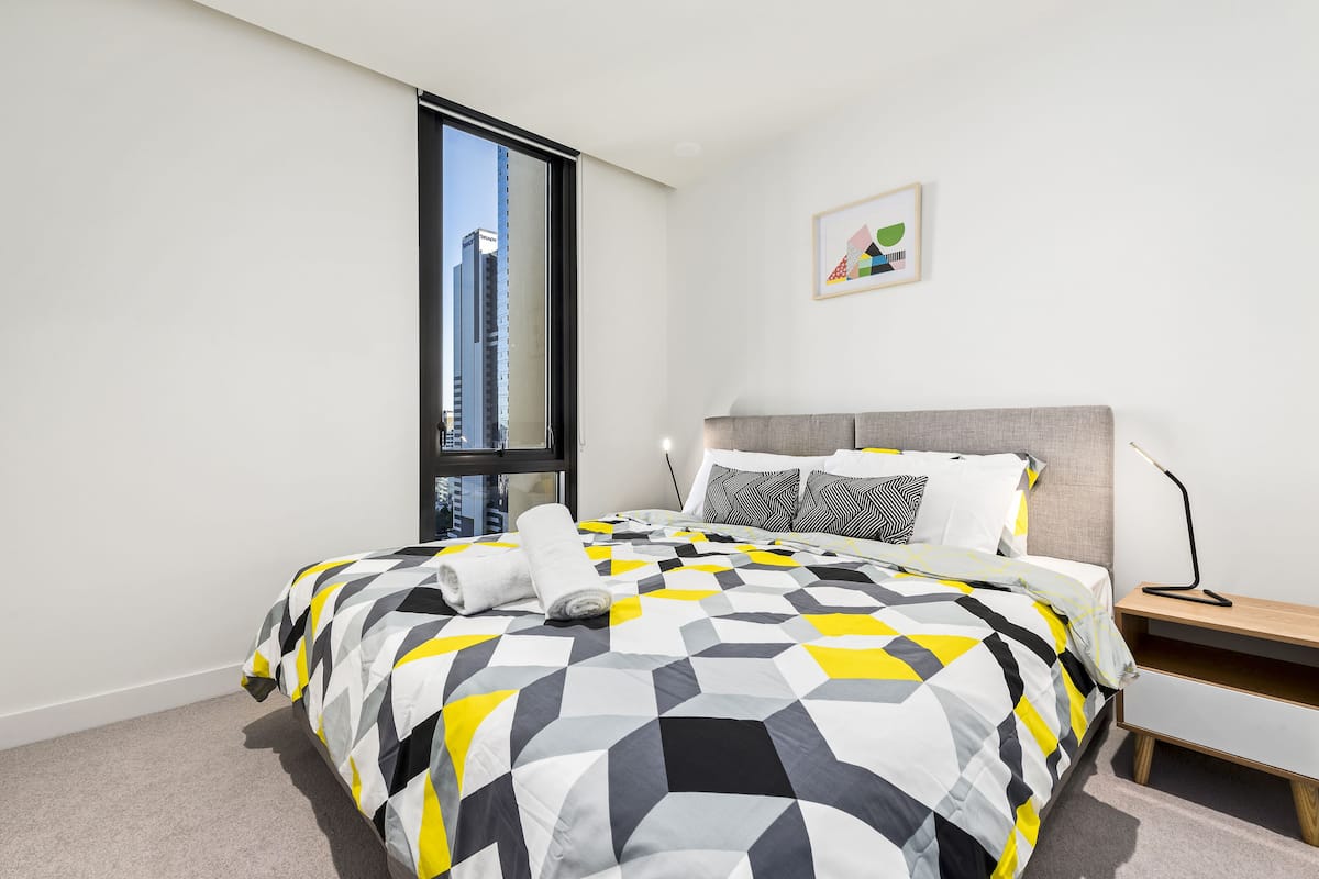 A modern hotel room with a large bed, a nightstand, a desk, a chair, and a window offering a city view.