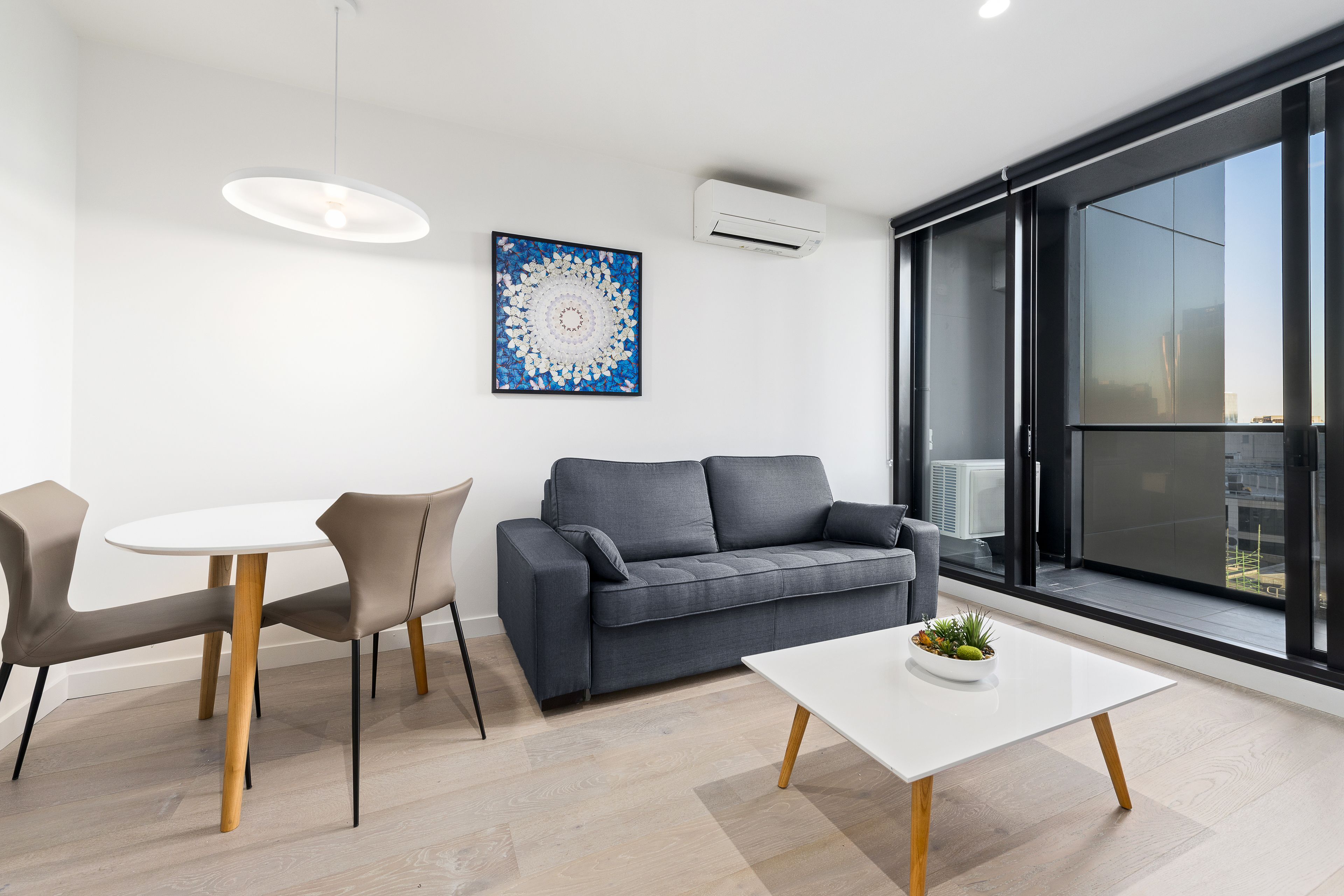 Un salon moderne avec un canapé gris, deux fauteuils, une table blanche et une grande fenêtre donnant sur un beau paysage.
