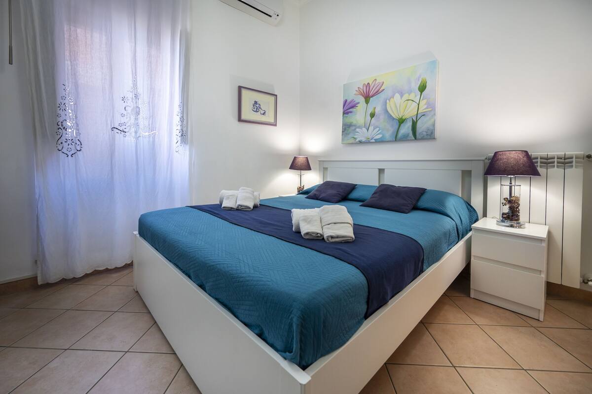 Ein ordentlich bezogenes Bett mit blau und dunkelblau gestreifter Bettwäsche, zwei Nachttische mit Lampen, ein Blumenbild an der Wand und ein Fenster mit durchscheinenden Vorhängen.