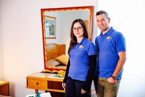 A man and a woman standing in a room with a wooden dresser, a mirror, and a bed with a floral arrangement.