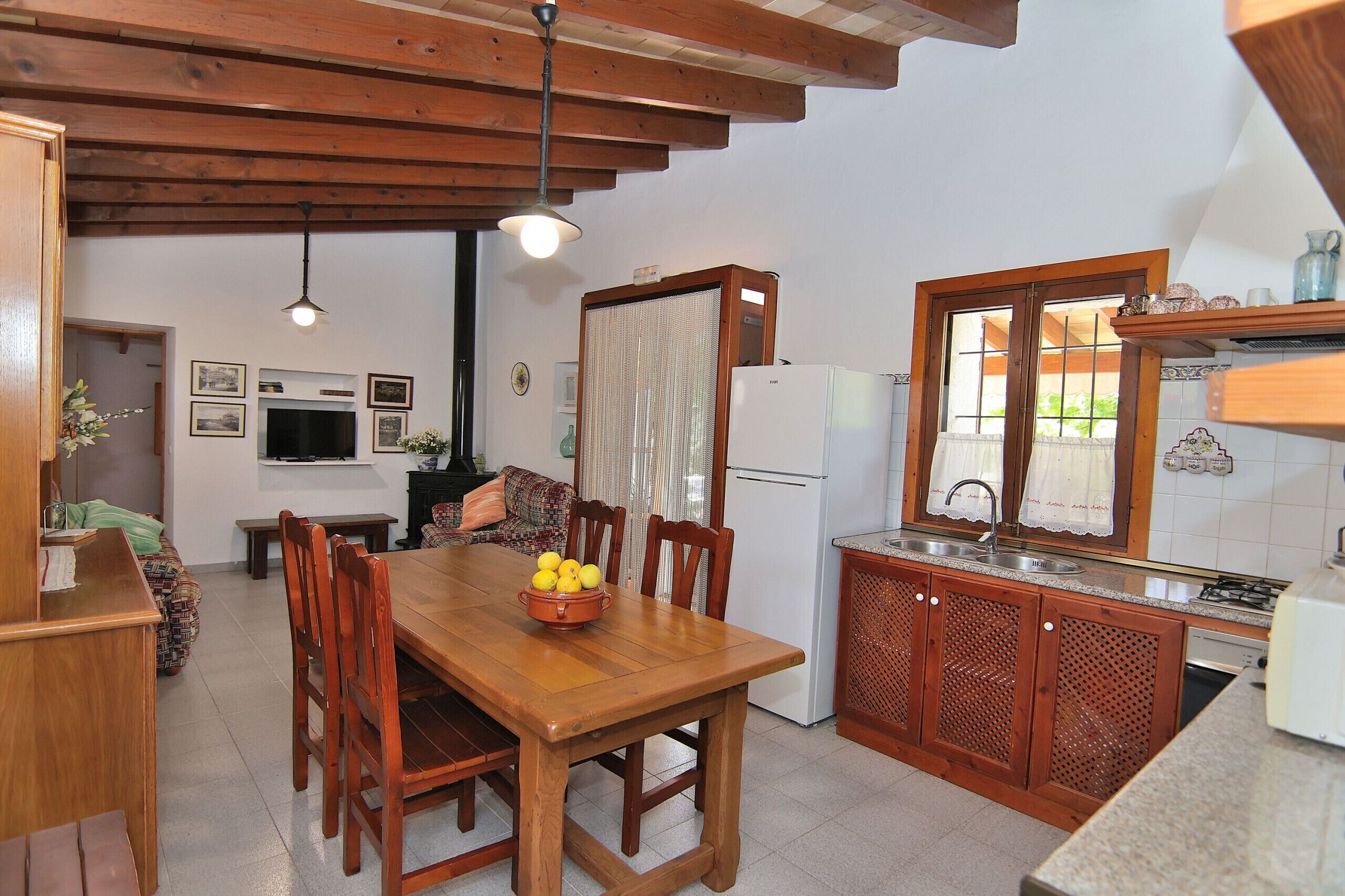 A compact kitchen with wooden cabinets, a dining table with chairs, a refrigerator, and a window with curtains.