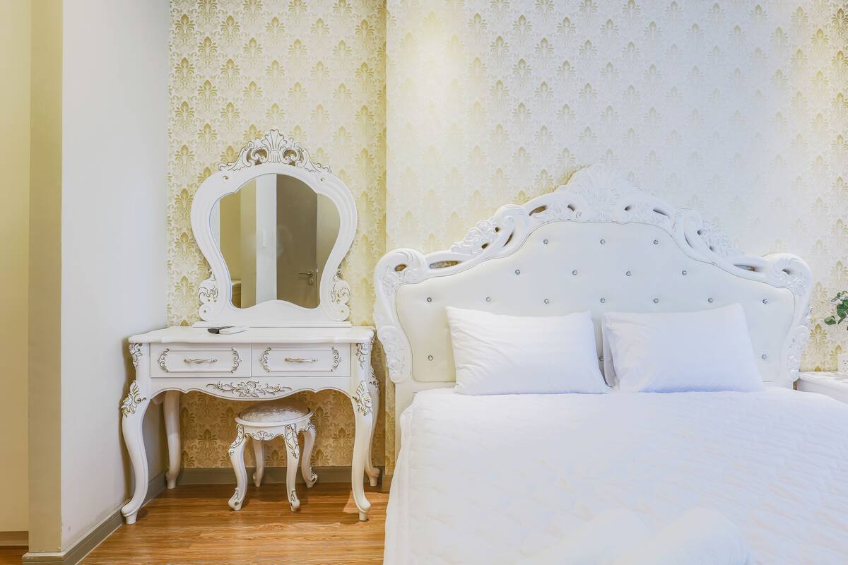 A bedroom with a white bed, a white vanity with a mirror, and a small stool.